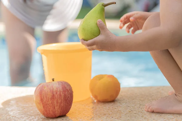 Jolie petite main de garçon tenant une poire fraîche. Un seau en plastique jaune et quelques fruits autour . — Photo