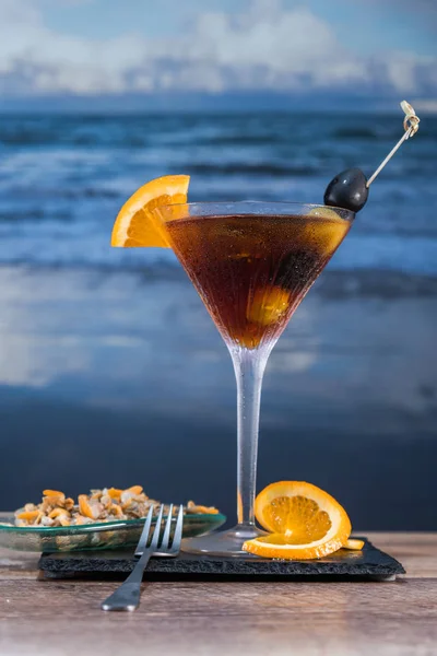 Coquetel vermelho com gelo, azeitonas, fatias de limão e conchas em uma mesa de vidro com ondas do mar no fundo - Azeitonas verdes e pretas — Fotografia de Stock
