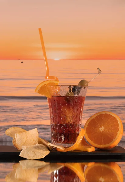 Экзотический коктейль с оливками, оранжевыми ломтиками и картошкой фри на фоне морских волн - на стеклянном столе — стоковое фото