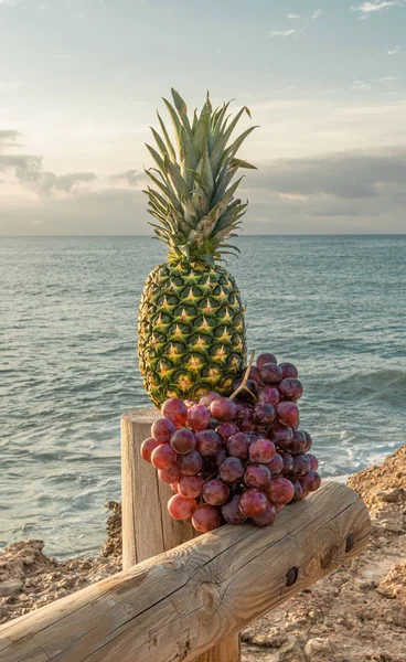 Heerlijke tropische snack-een volumineuze groene ananas vergezeld van een lommerrijke cluster van grote druiven op een houten steiger-de zee en de blauwe hemel op de achtergrond — Stockfoto
