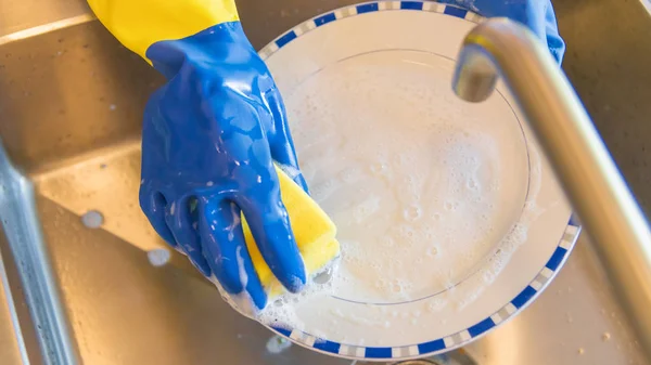 Wash the dishes in the kitchen - Blue and yellow plastic gloves with a lot of foam - Male washing the dishes in the kitchen sink area