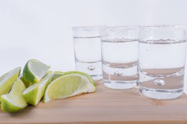 Ahşap kesme tahtası üzerine yığılmış yakın çekim limon dilimleri ve beyaz arka plan üzerine üç tekila bardağı