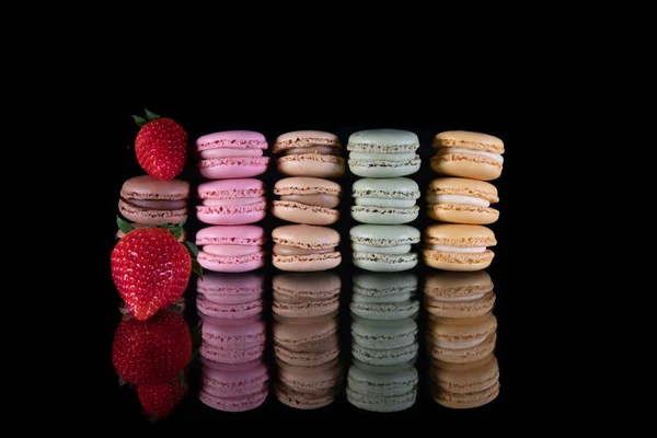 Dois suculentos morangos vermelhos ao lado de macaroons marrom, rosa, verde e amarelo empilhados em uma mesa de vidro com um fundo preto — Fotografia de Stock