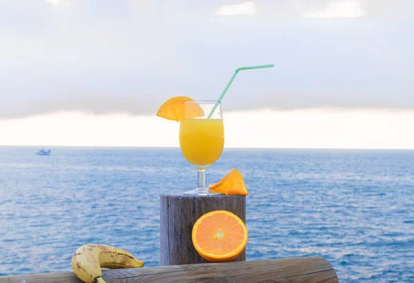 Fatias de laranja madura, banana e um delicioso suco de laranja Barcos no mar azul sob fundo de céu nublado — Fotografia de Stock