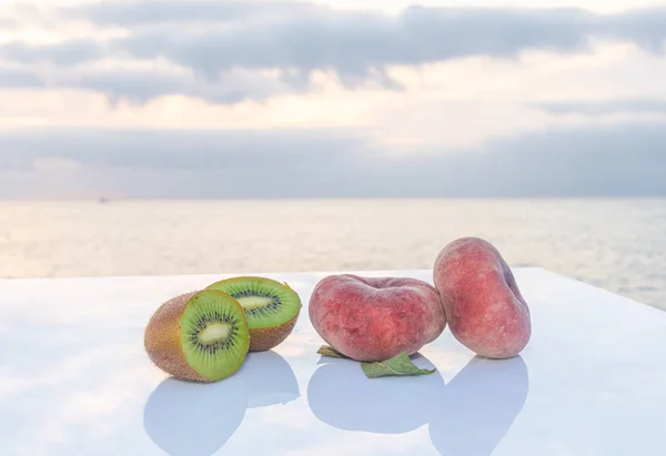 Два вкусных плоских персика в сопровождении свежего зеленого киви, разрезанного пополам на белом столе - Море и голубое небо на заднем плане — стоковое фото