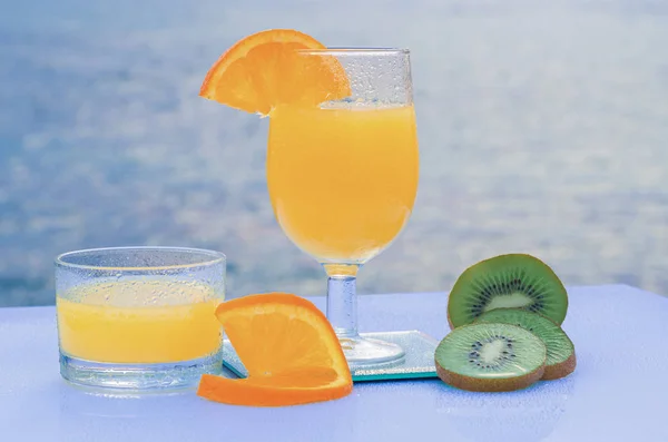 Green kiwi slices and orange juice for breakfast - The blue sea under a cloudy sky in the background — Stock Photo, Image