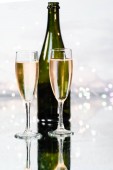 Dva luxusní skleněné šálky plněné žlutým šampaňským a zelenou láhev za pozadím osvětlená mnoha malými vánočními světly. Koncept večírku a oslavy.