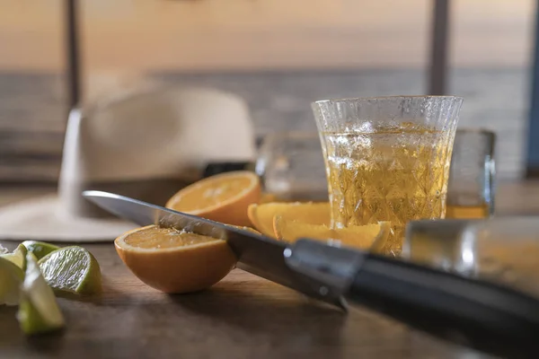 Нож режет апельсин, ломтики лимона и рюмку виски с кубиками льда, белая шляпа на заднем плане вне фокуса . — стоковое фото