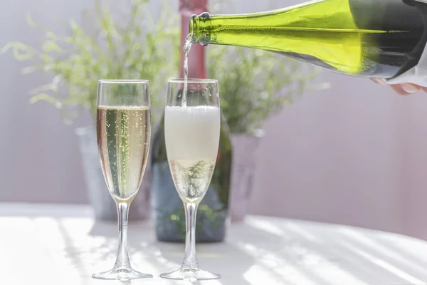 Um barman enchendo uma taça de champanhe ao lado de outra taça cheia de champanhe, uma garrafa verde de champanhe e um vaso no fundo . — Fotografia de Stock