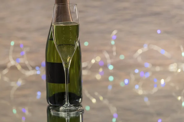 Una copa de cristal con un champán amarillo con una botella verde detrás en una mesa de cristal con muchas luces de Navidad en el fondo fuera de foco. Concepto de celebración . — Foto de Stock