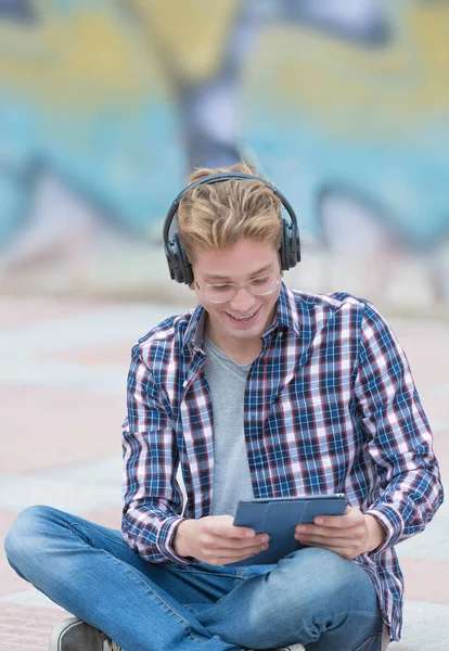 Niño rubio sonriente con auriculares, sentado en el suelo viendo vídeos en su tablet con una pared con graffiti en el fondo. Concepto de juventud tecnológica . —  Fotos de Stock