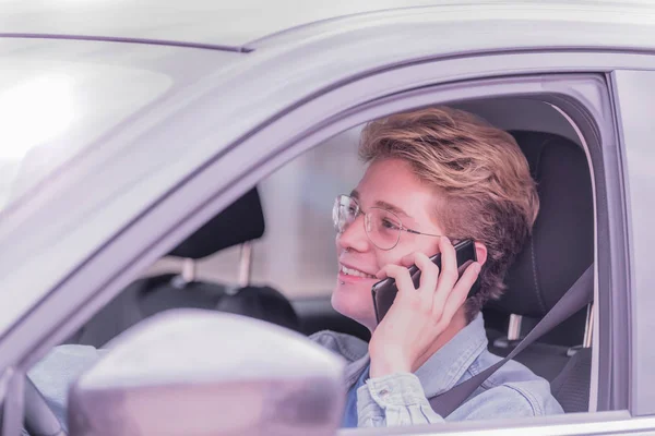 Menino loiro sentado em um carro e olhando em frente e sorrindo enquanto fala ao telefone . — Fotografia de Stock