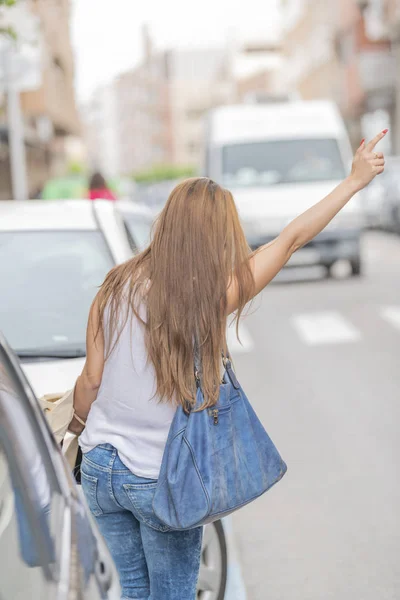 Vista trasera de la chica rubia levantando la mano como si quisiera detener un taxi con camisa blanca con pantalones azules, billetera azul con fondo borroso. Imagen vertical . — Foto de Stock