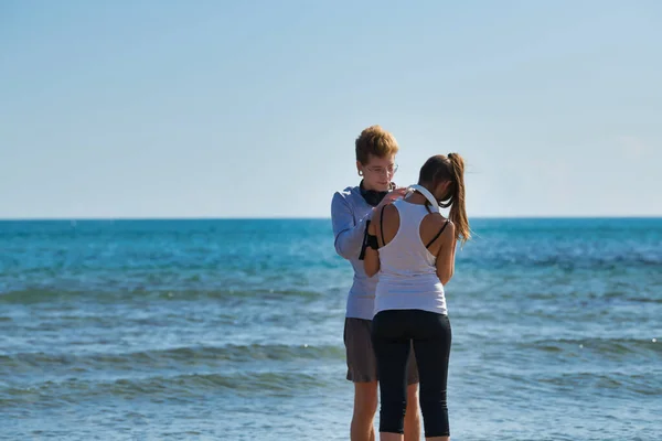 若いブロンドの男は、午前中に海の海岸を走っているときにガールフレンドのヘッドフォンに適合します。演習で健康的な生活の概念. — ストック写真
