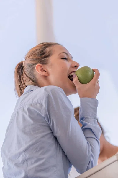 Schattige jonge vrouw draagt een lichtblauw shirt en verzamelde haar houdt een appel in een van haar handen waardoor het naar haar mond. — Stockfoto