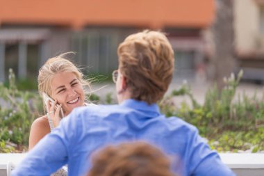 Güneşli bir günde kameraya geri dönen erkek arkadaşıyla aynı masada otururken telefonda gülümseyen kız..