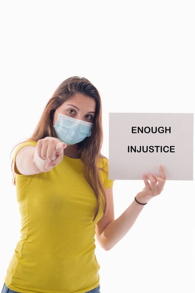 Young woman using a surgical mask holding a sign with a message and pointing at the camera on a light background. Racism and awareness concept.