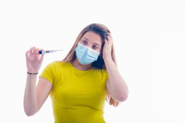 Sol eli başının üzerinde cerrahi maske kullanan genç bir kadın arka planda dijital termometreyi kontrol ediyor. Sağlık ve hastalık konsepti.