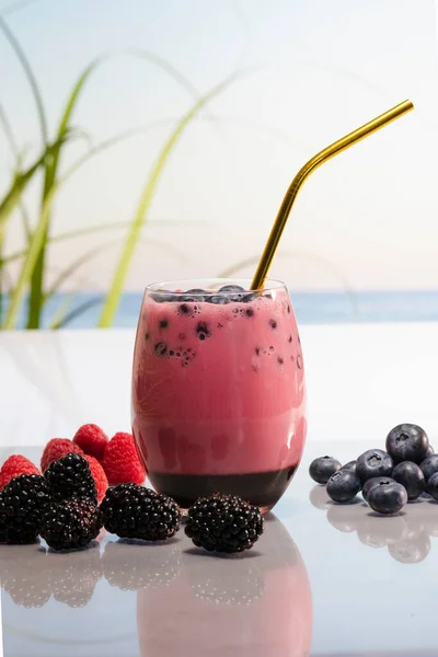 Close up of a delicious looking glass of fruit juice with blueberries and a straw surrounded by raspberries, blackberries and some more blueberries on an out of focus background. Holiday and healthy food concept.