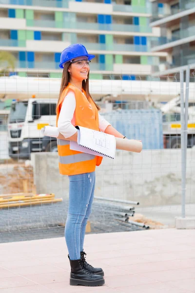 Smiling beautiful architecture student wearing safety gear and holding blueprints outside a construction site. Work and apprenticeship concept.