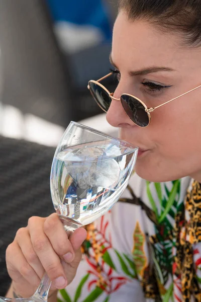 Close up of a beautiful woman wearing glasses drinking from a glass of water with ice cubes on it on an out of focus background. Healthy lifestyle and holidays concept.