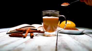 Tarçın çubuklarıyla çevrili bir fincan çaya kahverengi şeker, açık bir kitap ve ahşap bir masada bir limon ekleyen bir erkek eli. Rahatlama ve yaşam tarzı kavramı.
