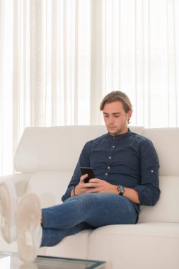 Kanepede oturan yakışıklı genç adam, ayakları masanın üstünde, arka planda telefonuna bakıyor. İletişim ve eğlence kavramı.