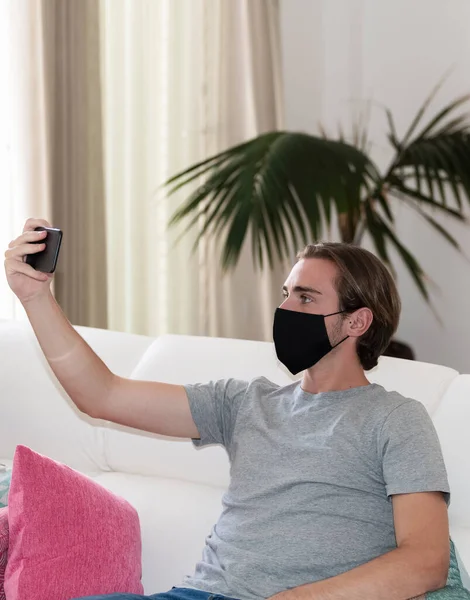 ソファに座っている顔のマスクを使用してハンサムな若い男は 彼の携帯電話を離れて保持し フォーカスの背景の外にそれを見て レジャーとコミュニケーションのコンセプト — ストック写真