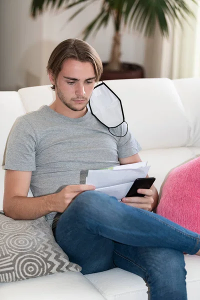 Handsome young man with a face mask hanging from his left ear checking some papers and holding his phone while sitting on a sofa. Payments and economy concept.
