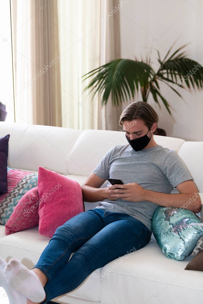 Handsome young man wearing a face mask checking his phone while sitting on a sofa on an out of focus background. Relaxation and communication concept.