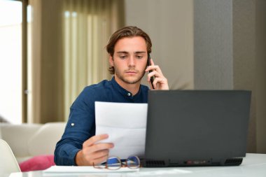 Yakışıklı bir genç, arka planda bir dizüstü bilgisayarın önünde telefonunu kullanırken faturayı kontrol ediyor. Ödemeler ve teknoloji kavramı.