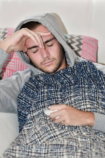 パーカーを着た若い男がナプキンを持ってソファの上の毛布の下で休んで閉じます 病気と医療の概念 — ストック写真