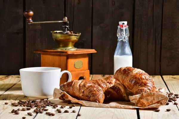 Appetizing croissants surrounded by coffee beans, a cup, a traditional coffee mill and a glass bottle of milk on a table. Breakfast concept.