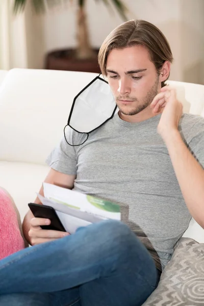 Handsome young man with a face mask hanging from his right ear reading some papers and holding his phone while sitting on a sofa on an out of focus background. Payments and economy concept.