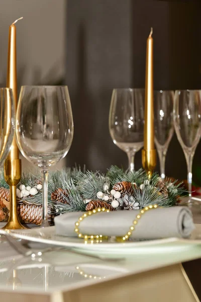 select focus of a pine-cone wreath surrounded by glasses and candles on a table on an out of focus background. Christmas holiday and celebration concept.