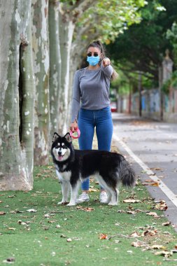 Gözlüklü ve cerrahi maskeli bir kadın arka planda kameraya çevrilmiş bir köpeği tasmayla tutuyor. Yeni normal ve evcil hayvan kavramı.