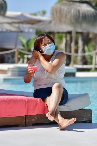 アジアの女性が飲み物を持っていて 集中力のない背景でホテルのテラスに座って手術用マスクを固定しています 新しい休日と安全コンセプト — ストック写真