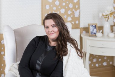 Güzel makyaj ve kar beyazı gülümseme, kıvırcık saçlı bir esmer ile curvy kadın. Bir koltuk üzerinde oturan bir ekose olarak, stüdyo fotoğrafı