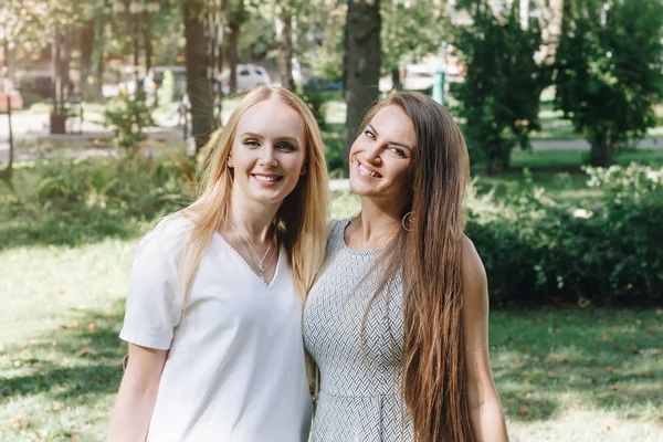 Two beautiful girls brunette and blonde, happy walk in the park, students sisters, girlfriends walk in the sunny summer outdoors. Smiling women, positive emotions