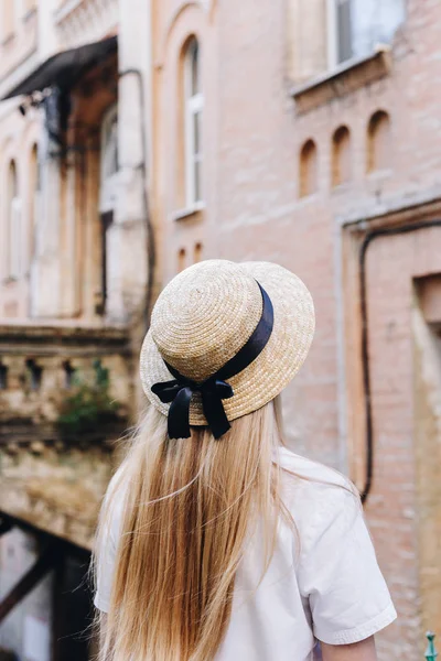 A thin blonde girl with long hair in a white dress stands with her back to the camera and looks into the distance in a wide-brimmed hat. City landscape, traveler.