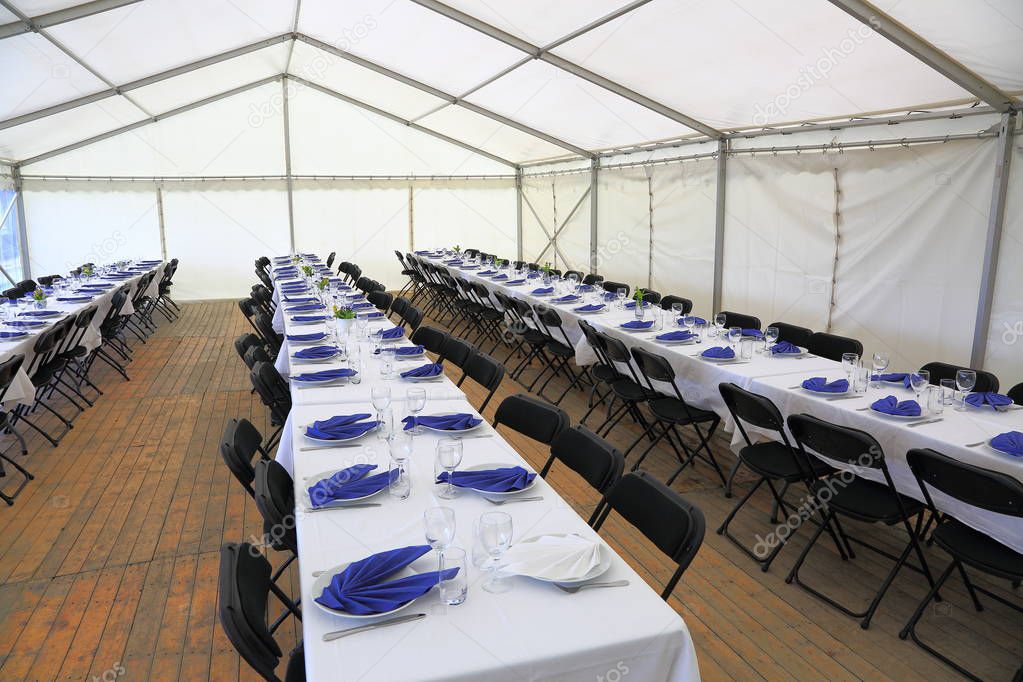 View of rented tent ready for guests. White table cloth, white plates with blue napkins and empty glass. Party celebration concept. 