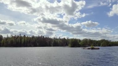 Kayalık kıyı şeridi, mavi su yüzeyi ve beyaz bulutlu mavi gökyüzünde yeşil ağaçlar. İsveç, 