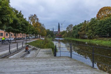 Güzel bir sonbahar şehri manzarası. Genç kadın silueti nehir kenarında oturuyor. Arka planda beyaz bulutlar olan mavi gökyüzü. 10.01.2020. Uppsala. İsveç