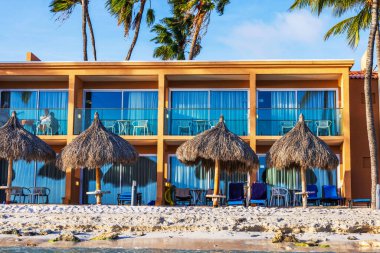 Karayip Denizi 'ndeki kumlu sahilde palmiye ağaçları ve saman şemsiyeleri olan sahil manzaralı oteli kapat..