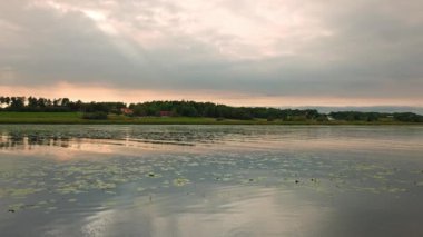 Sakin gölün üzerinde gün batımının güzel manzarası. Akşam güneşinin aydınlattığı bulutlu gökyüzü ve nilüferler. İsveç.