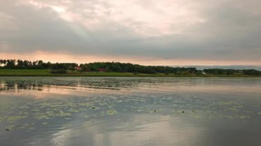 Sakin gölün üzerinde gün batımının güzel manzarası. Akşam güneşinin aydınlattığı bulutlu gökyüzü ve nilüferler. İsveç.