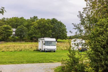 Ağaçların ve kırsal evin yakınındaki yeşil çayırda bulutlu arka planda park etmiş iki karavan. İsveç.