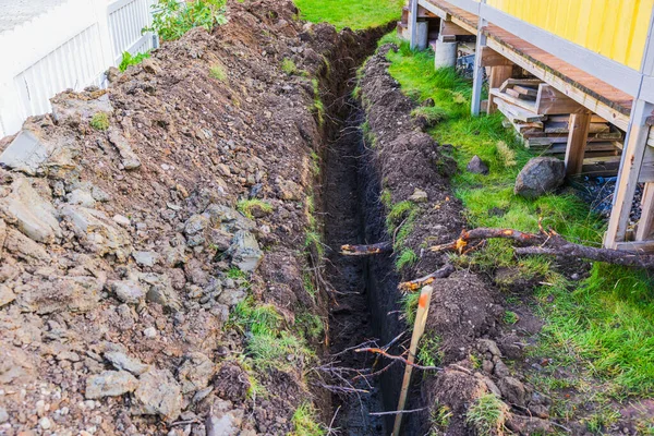Açıkta toprak, kök ve inşaat malzemeleri olan sarı ahşap evin yanında yeni kazılmış siper. İsveç.
