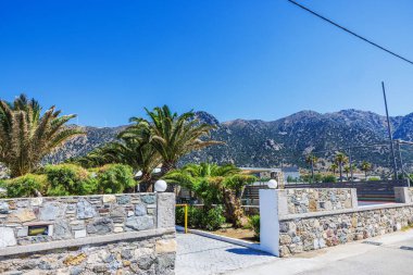 Uzun palmiye ağaçları ve açık mavi gökyüzü olan kayalık dağların arka planındaki taş duvar ve otele giriş. Yunanistan. Kos.