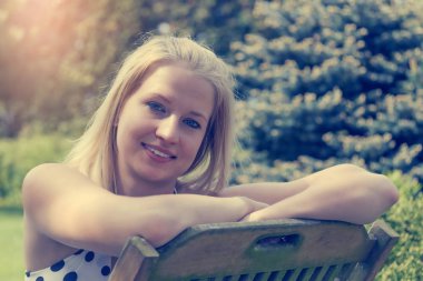 Bahçedeki tahta sandalyede oturan havalı, gülümseyen sarışın kadının portresi.. 